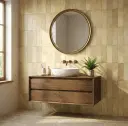 Bathroom interior featuring sandy beige patterned wall tiles, floating wooden cabinet, round mirror, white countertop basin, and small plant.
